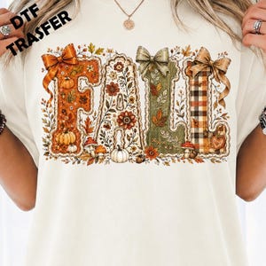 May include: A cream-coloured t-shirt featuring a graphic print of the word FALL in large, decorative letters. The letters are filled with autumn-themed illustrations including pumpkins, mushrooms, leaves, and flowers, accented with bows.