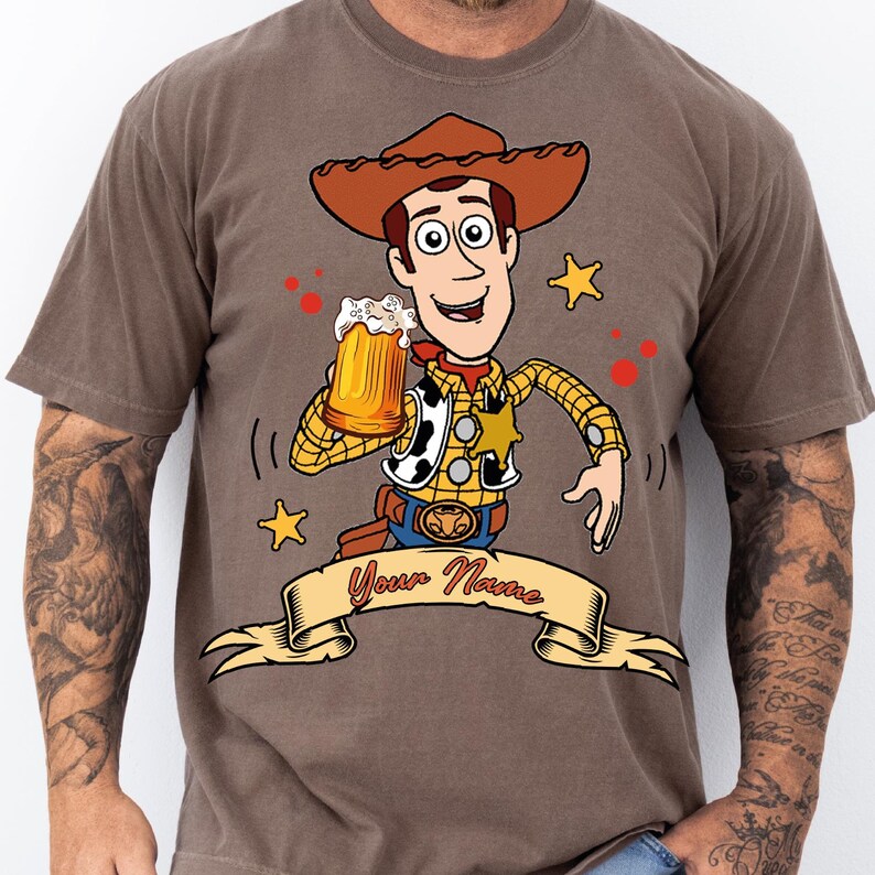 Może przedstawiać: Brązowy t-shirt z grafiką przedstawiającą postać kowboja z kresk&oacute;wki w kapeluszu, ż&oacute;łtej koszuli w kratę i kamizelce, trzymającego kufel pienistego piwa. Poniżej znajduje się beżowy baner z napisem Your Name wykonanym czerwoną kursywą.