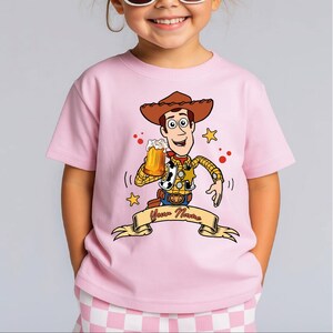 Może przedstawiać: Jasnor&oacute;żowy t-shirt z kr&oacute;tkim rękawem z grafiką przedstawiającą kowboja z kresk&oacute;wki w brązowym kapeluszu, ż&oacute;łtej koszuli w kratę i kamizelce w łaty, trzymającego kufel piwa, z banerem poniżej zawierającym napis Your Name.