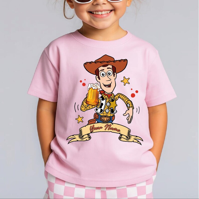 Może przedstawiać: Jasnor&oacute;żowy t-shirt z kr&oacute;tkim rękawem z grafiką przedstawiającą kowboja z kresk&oacute;wki w brązowym kapeluszu, ż&oacute;łtej koszuli w kratę i kamizelce w łaty, trzymającego kufel piwa, z banerem poniżej zawierającym napis Your Name.
