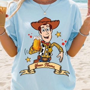 Może przedstawiać: Jasnoniebieski t-shirt z grafiką przedstawiającą postać kowboja z kresk&oacute;wki w brązowym kapeluszu, ż&oacute;łtej koszuli w kratę i kamizelce, trzymającego kufel piwa. Pod postacią znajduje się beżowy baner z napisem Your Name wykonanym czerwoną czcionką.