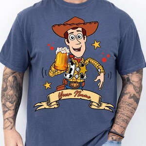 Może przedstawiać: Niebieski t-shirt z grafiką przedstawiającą postać kowboja w brązowym kapeluszu, ż&oacute;łtej koszuli w kratę i kamizelce w łaty. Postać trzyma kufel piwa. Poniżej znajduje się beżowy baner z czerwonym napisem Your Name.