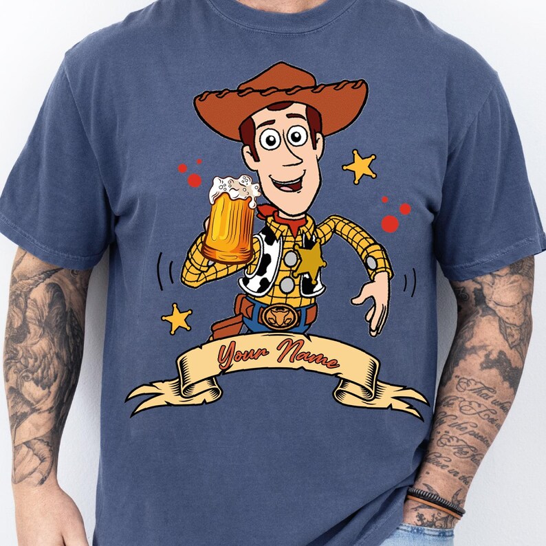 Może przedstawiać: Niebieski t-shirt z grafiką przedstawiającą postać kowboja w brązowym kapeluszu, ż&oacute;łtej koszuli w kratę i kamizelce w łaty. Postać trzyma kufel piwa. Poniżej znajduje się beżowy baner z czerwonym napisem Your Name.