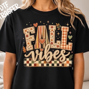 May include: A black short-sleeved t-shirt featuring the text FALL vibes in a decorative font. The letters are filled with floral, striped, and plaid patterns in orange, brown, and cream. Small hearts, stars, and a checkered border are included.