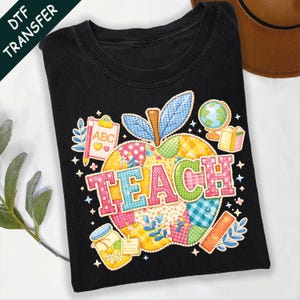 May include: Black t-shirt featuring a colourful apple design. The apple is made of patchwork fabric and the word "TEACH" is across the front. The design includes school-themed elements like a globe, ruler, and jar of honey.