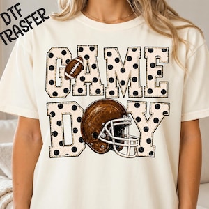 May include: A cream-colored t-shirt featuring the text GAME DAY in large block letters with a black polka dot pattern. A brown football replaces the letter A, and a brown football helmet with a white face mask is positioned over the letter D.