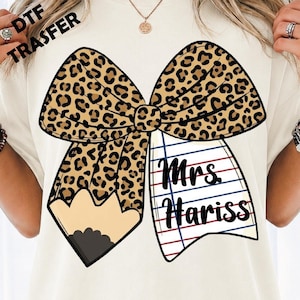 May include: A cream-colored t-shirt featuring a large graphic of a bow. The bow has a leopard print pattern on one side and a white lined notebook paper pattern on the other. The text "Mrs. Hariss" is printed in black on the notebook section.