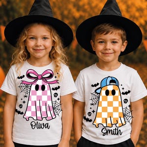 May include: Two white short-sleeve t-shirts featuring ghost graphics. One ghost has a pink checkered pattern and a pink bow, labelled 'Oliva'. The other ghost has an orange checkered pattern and a blue baseball cap, labelled 'Smith'. Both include spiderwebs, bats, and skulls.