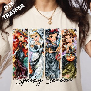 Peut inclure: Un t-shirt couleur crème avec une impression graphique de quatre princesses de dessin animé en tenue d'Halloween. Le texte "DTF TRASFER" apparaît dans le coin supérieur gauche et "Spooky Season" est écrit en lettres cursives noires en bas.