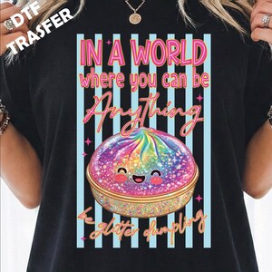 May include: A black t-shirt featuring a graphic of a rainbow-colored, glittery dumpling with a smiling face. The text reads, "IN A WORLD where you can be Anything be glitter dumpling," set against a background of vertical light blue and white stripes.
