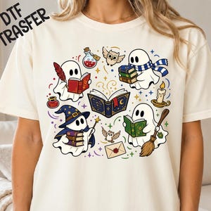 May include: A cream-coloured t-shirt featuring a graphic print of four white ghosts reading books, holding a broom, wearing a wizard hat, and surrounded by small owls, a candle, a potion bottle, an inkwell, and a sealed envelope with stars.