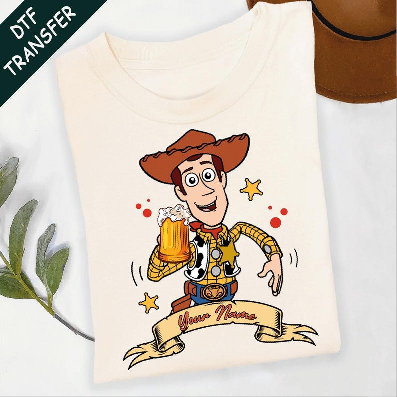 Może przedstawiać: Kremowy t-shirt z rysunkowym kowbojem w brązowym kapeluszu, ż&oacute;łtej koszuli w kratę i kamizelce w łaty. Postać trzyma kufel piwa, otoczona ż&oacute;łtymi gwiazdkami i czerwonymi kropkami, z banerem poniżej z napisem Your Name na dole grafiki.