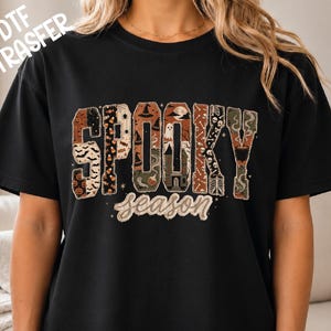 May include: A black t-shirt featuring a large graphic with the word SPOOKY in block letters filled with various Halloween patterns including pumpkins, bats, ghosts, and a haunted house, with the word season written below in a cursive font.