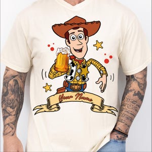 Może przedstawiać: Kremowy t-shirt z rysunkowym kowbojem w brązowym kapeluszu, ż&oacute;łtej koszuli w kratę i kamizelce w łaty. Postać trzyma kufel piwa nad beżowym banerem z czerwonym napisem Your Name.