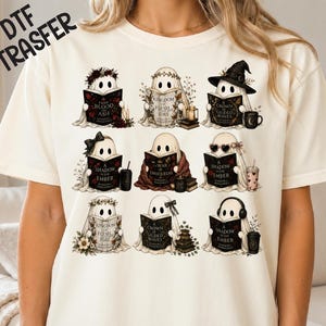 May include: A cream-coloured t-shirt featuring a graphic print of nine ghosts reading books. Each ghost is depicted with different accessories like flower crowns, hats, glasses, or headphones, alongside book titles such as From Blood and Ash.