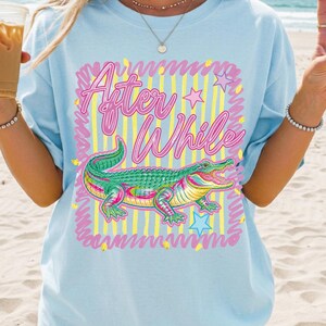 May include: Light blue t-shirt featuring a colorful alligator graphic with the text "After While". The design includes pink, yellow, and green accents, with stars and a striped background. A casual, relaxed fit tee.