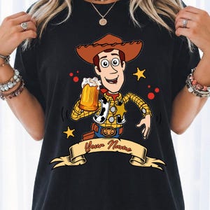 Może przedstawiać: Czarny t-shirt z grafiką przedstawiającą postać kowboja z kresk&oacute;wki w brązowym kapeluszu, ż&oacute;łtej koszuli w kratę i kamizelce w łaty. Postać trzyma kufel piwa. Poniżej znajduje się beżowy baner z napisem Your Name.