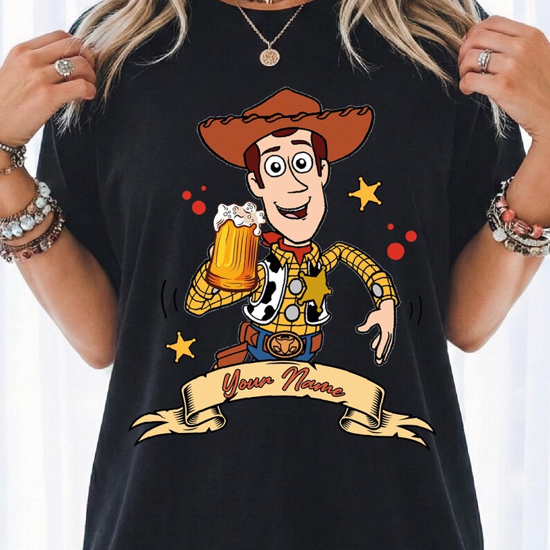 Może przedstawiać: Czarny t-shirt z grafiką przedstawiającą postać kowboja z kresk&oacute;wki w brązowym kapeluszu, ż&oacute;łtej koszuli w kratę i kamizelce w łaty. Postać trzyma kufel piwa. Poniżej znajduje się beżowy baner z napisem Your Name.