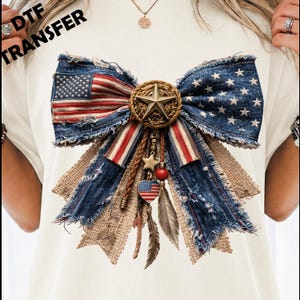 May include: A cream-colored t-shirt featuring a large graphic of a frayed denim bow with an American flag pattern. The center of the bow has a gold star on a textured circle, with dangling charms including a small star, a red bead, and a heart.