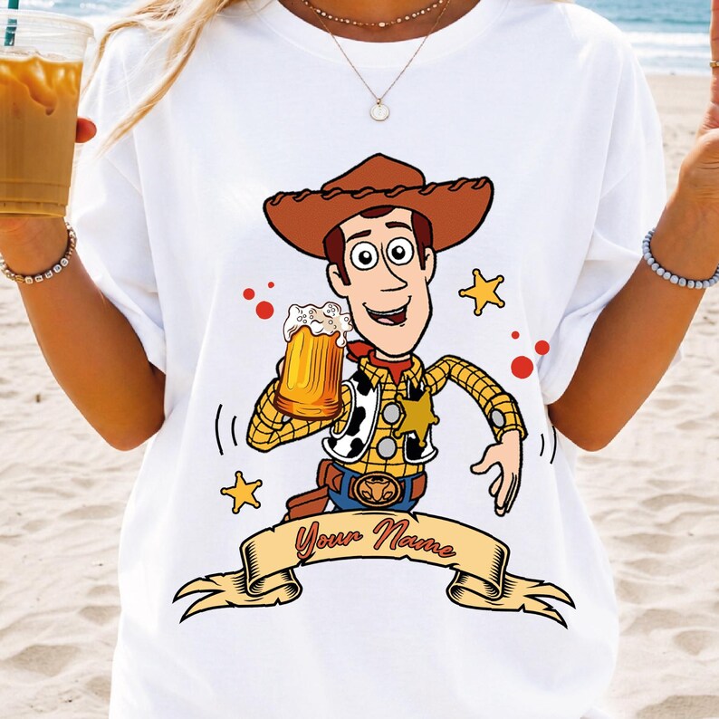 Może przedstawiać: Biały t-shirt z grafiką przedstawiającą postać kowboja w brązowym kapeluszu, ż&oacute;łtej koszuli w kratę i kamizelce w łaty. Postać trzyma kufel piwa, otoczona ż&oacute;łtymi gwiazdkami i czerwonymi kropkami, z banerem z napisem Your Name.