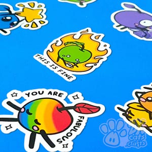 Könnte beinhalten: Eine Sammlung farbenfroher Aufkleber mit Cartoon-Obst- und Charakterdesigns. Ein Aufkleber zeigt einen Regenbogenapfel mit dem Text "YOU ARE FABULOUS". Ein anderer zeigt einen grünen Apfel in Flammen mit dem Text "THIS IS FINE."