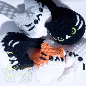 May include: A collection of handmade crochet cat toys in various colors. The cats are white, black, gray, and orange, with black and green eyes. The toys are soft and fluffy, with detailed facial features. The logo "cats crafts" is visible.