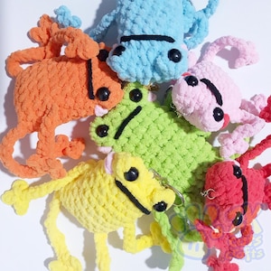 May include: A collection of colorful, handmade crochet frog keychains in various shades including orange, blue, pink, green, yellow, and red. Each frog has black eyes and a stitched mouth. The keychains are soft and plush.