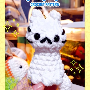 May include: A white crochet cat keychain with black eyes and a mustache. The keychain is handmade and features the text "Sitty Kitty Keychain Crochet Pattern" in a blue font. The website address ZKCRAFTCRAFTS.COM is also visible.