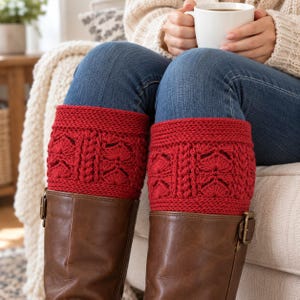 May include: A pair of bright red knit boot cuffs featuring a textured cable and fan stitch pattern. The cuffs are worn over blue denim jeans and tucked into brown leather boots with side buckles, providing a decorative layered appearance.