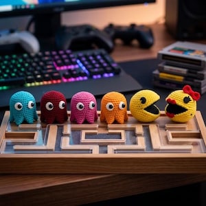 May include: A set of six crocheted figures including four ghosts in turquoise, maroon, pink, and orange, plus two yellow circular characters, one with a red bow, all arranged on a wooden maze board with clear acrylic dividers and a gray base.