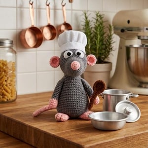 Puede incluir: Una figura de rata tejida a ganchillo de color gris con un gorro de chef blanco, orejas, nariz y patas rosas. La rata sostiene una pequeña cuchara de madera tejida a ganchillo marrón. Junto a ella hay una olla de metal con tapa y un cuenco.