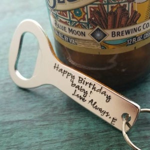 May include: A silver bottle opener with a key ring, resting on a teal surface. The opener has the text "Happy Birthday baby! Love Always. E" engraved on it. A partially visible Blue Moon beer bottle is in the background.