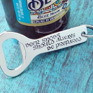 Può includere: Un apribottiglie d'argento con un portachiavi attaccato. L'apribottiglie ha inciso il testo "Beer Snobs Should Always Be Prepared".