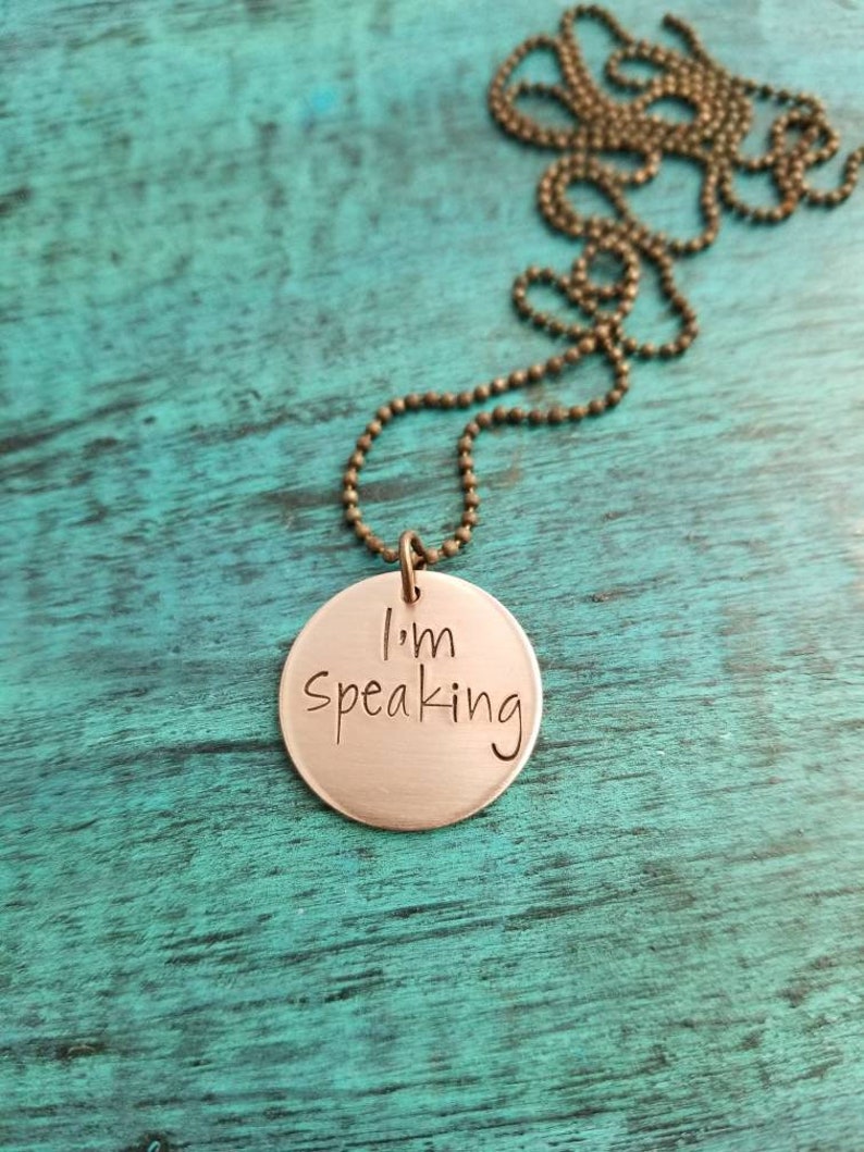 Pu&ograve; includere: Una collana di rame con un ciondolo rotondo che dice "Sto parlando". La collana &egrave; su una superficie di legno blu-verde.