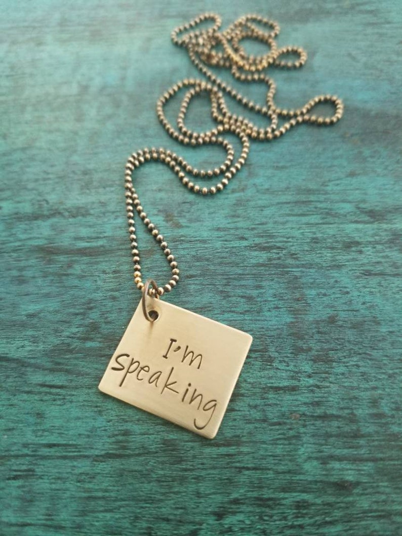 Pu&ograve; includere: Una collana d'oro con un ciondolo quadrato che dice "Sto parlando" in scrittura a mano.