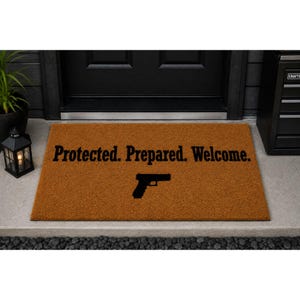 May include: A rectangular brown coir doormat featuring the black text "Protected. Prepared. Welcome." printed in a serif font. Below the text, a black silhouette of a semi-automatic handgun is centered on the mat's textured surface.