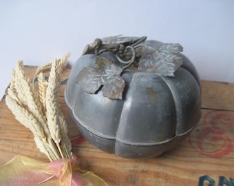 May include: A metallic, gray, pumpkin-shaped container with a smooth, segmented body and a decorative lid featuring sculpted metal leaves and a vine stem. Beside the pumpkin sits a small bundle of dried wheat stalks tied with a sheer ribbon.