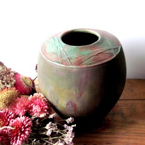 May include: A handcrafted ceramic vase with a wide, rounded body and a dark opening. The vase features a blend of green, brown, and red hues. The top rim has a textured design. Dried flowers in shades of pink and purple are arranged nearby.