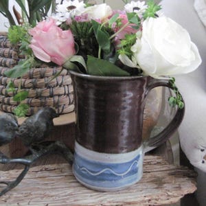May include: A brown ceramic mug with a blue and white glaze design. The mug is filled with a bouquet of pink and white flowers.