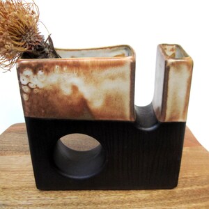 May include: A ceramic tape dispenser with a unique design. The top section is a blend of brown and cream, while the base is black. A dried flower is placed in the dispenser. The dispenser is sitting on a wooden surface.