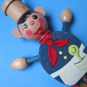May include: Wooden chef doll with a light brown head and hat, black hair, and rosy cheeks. The doll wears a blue and white uniform with a red neckerchief. The arms and legs are jointed, and the doll is set against a blue background.