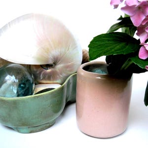 May include: A sage green ceramic bowl containing a large iridescent white seashell and a dark glass sphere, positioned next to a cylindrical light pink ceramic vase holding a cluster of pink hydrangea flowers with green leaves.