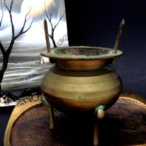 May include: A brass incense burner with a rounded body, three legs, and two upright handles extending from the rim. The metal surface shows a weathered patina with green oxidation near the base. The burner sits on a matching brass tray.