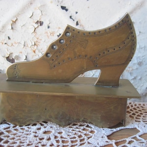 Può includere: Una scatola portaoggetti in ottone a forma di scarpa con un design ornamentale. La scatola è chiusa e ha un coperchio con cerniera. La scarpa è su una base con un design decorativo.