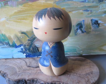 May include: A wooden Kokeshi doll with a round head and a cylindrical body. The doll has closed eyes, a small red mouth, and black hair. It wears a blue kimono with a floral pattern and a brown bow. The doll is displayed on a wooden slice.