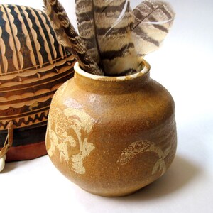 May include: A small, round, tan ceramic vase with a textured surface and light-colored floral patterns. Several brown and white striped feathers are placed inside the opening. Next to the vase is a rounded, tan and black patterned object.