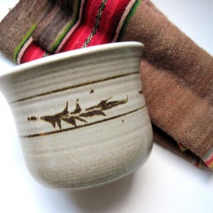 May include: A handmade ceramic bowl with a light gray finish and brown leaf design. The bowl is next to a folded brown and red patterned textile. The bowl is approximately 4 inches in diameter and 3 inches tall.