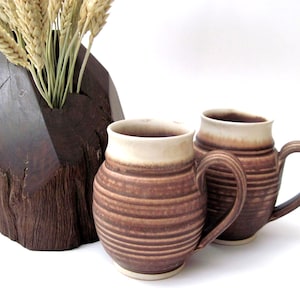 May include: Two brown ceramic mugs with cream-colored rims and handles sit next to a dark wooden vase holding wheat stalks. The mugs have horizontal striped detailing. The vase has a geometric shape and a natural wood grain.