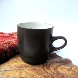 May include: A dark brown ceramic mug with a smooth, matte exterior finish and a contrasting white interior. The mug features a rounded handle on the right side and a slightly tapered base, presenting a simple, functional design for holding beverages.