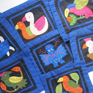 May include: A blue fabric patchwork quilt with black squares featuring colorful embroidered birds and other creatures. The quilt is made up of nine squares, each with a different design.